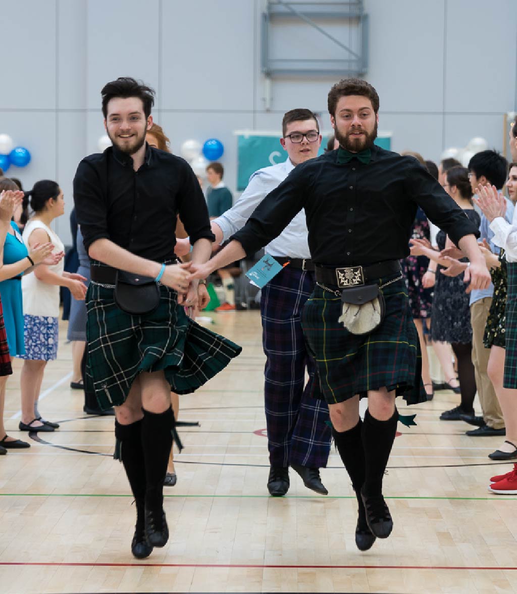 Three people wearing kilts dance forward in unison in a sports hall, with an audience standing on both sides clapping and watching.