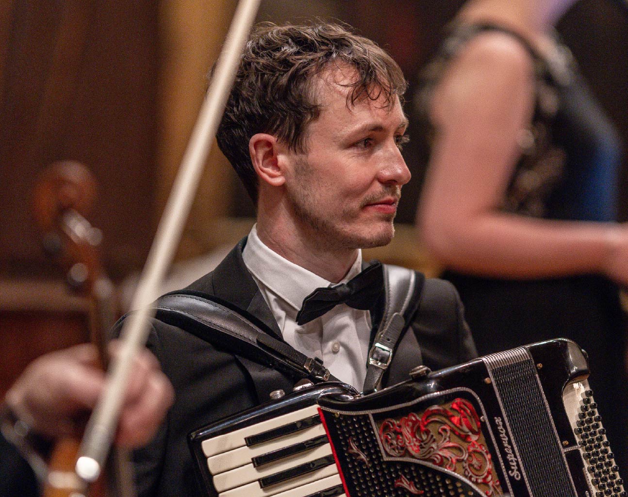 A musician in formal attire, including a black jacket and bow tie, plays an accordion during a live indoor performance. The accordion, decorated with red detailing and keys, is held across the front with straps. A violin and bow appear blurred in the foreground, while another performer and warm-toned wooden paneling are visible in the softly lit background.