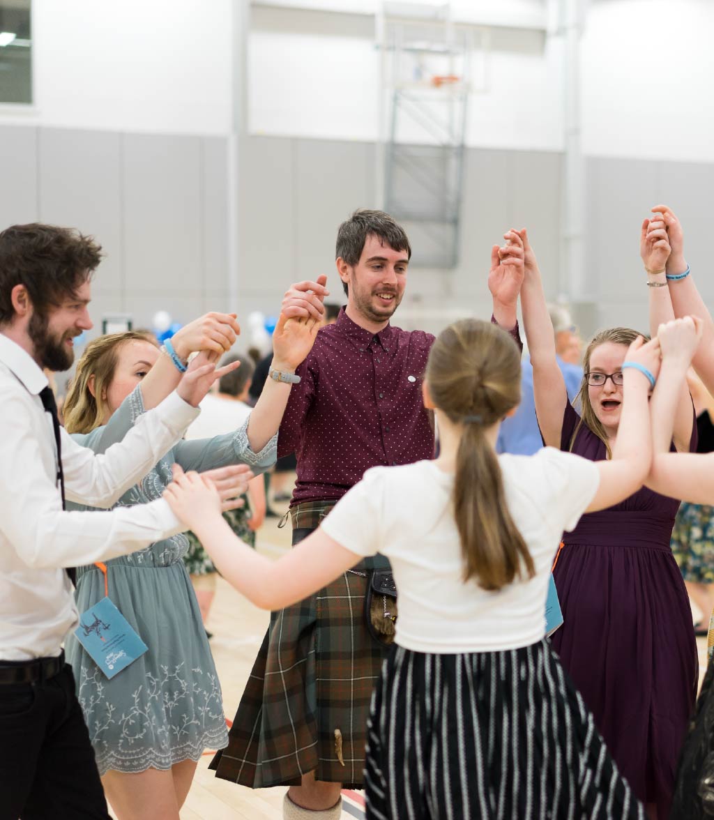 A group of people holding hands and raising their arms while dancing in a circle inside a large indoor hall. The participants wear a mix of formal and semi-formal clothing, including a tartan kilt, dresses, shirts, and skirts. The floor is a sports-style court, and a basketball hoop and light-colored walls are visible in the background, indicating a community or gymnasium setting.